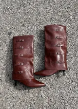 Botas altas de ecocuero color marrón con punta fina, taco kitten y múltiples tiras con hebillas metálicas plateadas a lo largo de la caña ancha.