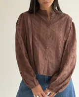 Blusa de brodery color blanco, con cuello redondo, mangas largas abullonadas con puños elásticos y cierre frontal con botones forrados. Presenta detalles de puntillas verticales en el frente.