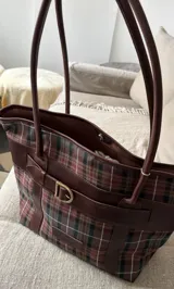 Cartera de mano tipo tote en diseño a cuadros escoceses en tonos verde, bordó y marrón, con detalles y asas en cuero marrón. Presenta una tira decorativa con hebilla metálica dorada en el frente.