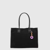 Tote bag de gamuza negra con asas largas de cuero sintético. Incluye un charm decorativo con cordón y una pieza circular de color lila con el logo grabado.