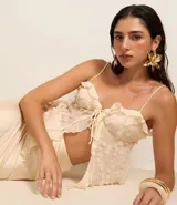 Blusa corta color crema con escote corazón, confeccionada en tejido con textura de lunares y volados de chifón. Presenta breteles finos ajustables y un lazo decorativo en el centro del busto.
