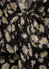 Blusa de gasa con estampado floral de margaritas sobre fondo negro, diseño recto semitransparente con cuello pico y detalle de lazo frontal. Cuenta con mangas largas y forro interior.