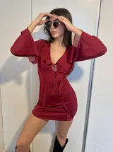 Vestido corto bordó ajustado al cuerpo, con escote en V pronunciado, volados en el cuello y lazo ajustable al frente. Presenta mangas largas de tul acampanadas.