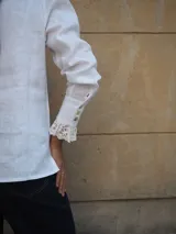 Camisa de lino color beige, con cuello bobo y detalles de encaje bordado en el cuello, pechera y puños. Cuenta con mangas largas levemente abuchonadas y cierre frontal con botones nacarados.