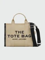 Bolso tipo tote de lona color negro con letras blancas bordadas en el frente que dicen 'THE TOTE BAG MARC JACOBS'. Cuenta con asas superiores cortas y una correa de hombro ajustable y extraíble.