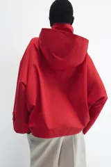 Cazadora roja de cuello subido con capucha y manga larga, bolsillos delanteros y cierre frontal cruzado con botones a presión dorados.