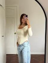 Polera de punto acanalado color blanco, con escote redondo y detalle de tiras cruzadas en el pecho, de manga larga y corte ajustado con ruedo curvo.