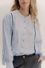 Camisa de algodón blanca con cuello a la base, cierre frontal con botones y mangas largas con puños delicados. Presenta volados verticales y detalles de cintas de terciopelo en contraste a lo largo del frente.