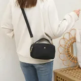 Cartera bandolera negra de lona, con cierre de cremallera, herrajes dorados y correa ajustable.