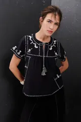 Blusa negra de algodón con bordados blancos a contratono, cuello redondo con lazo y borlas decorativas, y mangas cortas.