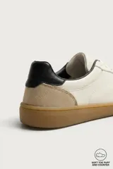 Zapatillas deportivas barefoot de hombre elaboradas en piel. Diseño en color blanco con detalles en negro y beige en el talón, y suela plana de goma marrón.