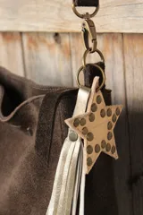 Bandolera confeccionada en cuero de gamuza marrón, con cierre superior. Presenta un llavero colgante con forma de estrella revestido en tachas metálicas y tiras flecadas en tono dorado.