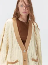 Cardigan de punto trenzado en color negro con detalles en color crema en los bolsillos, puños y ruedo. Presenta un diseño oversize, cierre frontal con botones y bordado decorativo en uno de los bolsillos delanteros.