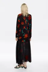 Vestido midi de corte ajustado con falda superpuesta de tul, escote barco y mangas largas tipo murciélago. Presenta un diseño estampado con motivos florales en tonos rojos y verdes sobre fondo negro, con detalles fruncidos en la cintura.