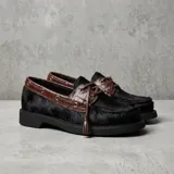 Mocasín de estilo náutico confeccionado en cuero, combinando una base de cuero con textura de pelo en color negro y detalles de cuero con textura croco en color marrón. Presenta cordones decorativos con borlas laterales y suela plana de goma.