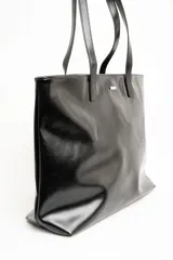 Bolso tipo tote bag color marrón, confeccionado en cuero sintético, con doble asa superior y detalle de placa metálica dorada con logo en la parte frontal.