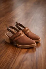 Zueco de cuero vacuno color suela, con diseño de tira trenzada y textura grabada en el empeine. Cuenta con una tira trasera ajustable con hebilla metálica, taco de madera y suela con plataforma baja.