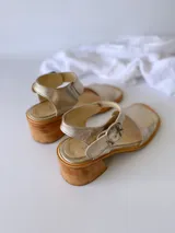 Sandalias de cuero color dorado, con tira ancha sobre los dedos, pulsera al tobillo con hebilla metálica y taco de madera.