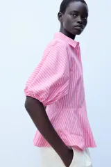 Camisa de popelín a rayas rosadas y blancas, con cuello solapa, mangas abullonadas por debajo del codo con terminación elástica, cintura entallada y bajo en evasé. Cierre frontal con botones ocultos.