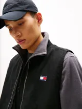 Chaleco negro de lona de algodón con cuello de punto canalé y cierre de cremallera. Tiene dos bolsillos ribeteados delanteros, un bolsillo interior de parche y parche de Tommy en el pecho.