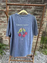 Remera azul con efecto lavado, de corte holgado y manga corta. Estampa trasera con la frase "Cherry Season" y un diseño pixelado de cerezas.