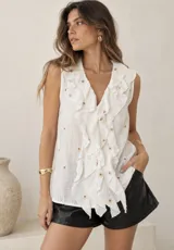 Blusa sin mangas de color beige, con escote en V pronunciado y grandes volados frontales. Presenta pequeños detalles bordados en toda la prenda.