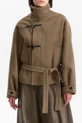 Chaqueta corta de silueta amplia confeccionada en mezcla de lana, poliéster y acrílico. Presenta cuello alto estructurado, cierre frontal con alamares y cinto ajustable que define la cintura. Incluye mangas largas con trabas abotonadas en los puños.