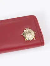 Billetera compacta de cuero vacuno color bordó, con cierre perimetral, correa de muñeca y aplique metálico dorado en forma de mariquita en el frente.