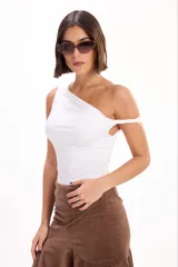 Blusa corta blanca de diseño asimétrico con un hombro descubierto y detalle de tiras cruzadas, confeccionada en tela liviana con fruncidos laterales.