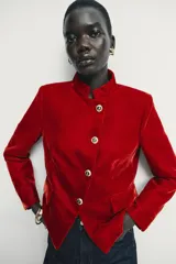 Blazer de terciopelo color rojo, con cuello subido, manga larga y cierre frontal cruzado con botones dorados. Presenta bolsillos delanteros con solapa.