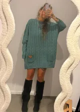 Sweater de punto color camel con diseño de trenzas verticales y acabado brillante. Presenta una silueta holgada tipo poncho, cuello redondo, mangas largas con puños ajustados y bolsillos frontales con detalle de etiqueta de cuero.