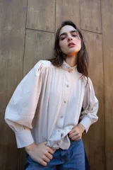 Camisa de algodón color crudo con cuello alto con volados, cierre frontal de botones, mangas largas abullonadas con puños estructurados y detalle de frunces en los hombros.
