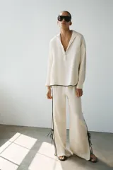 Camisa de lino color crudo con cuello solapa, manga larga y bajo asimétrico. Presenta detalles de ribetes negros a contraste y aberturas laterales con tiras para ajustar.