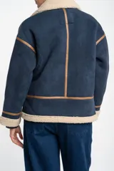 Campera tipo gamulán de corte oversize, color azul marino con interior y cuello de corderito en tono beige. Presenta detalles de costuras y bordes en cuero sintético color marrón, cierre frontal y mangas largas.