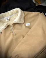 Buzo de pana color beige con cuello polo, cierre frontal parcial y forro interior de corderito. Presenta diseño oversized, bolsillos laterales y puños elásticos.