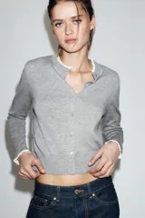 Chaqueta de punto de corte clásico, color gris, con cuello redondo y manga larga. Presenta cierre frontal con botones y detalles de puntilla en color crema en el cuello y los puños.