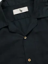 Camisa negra de manga corta con cuello camp y calce relajado. Confeccionada en una mezcla de lino y algodón, terminada con botones de madera y parche característico de Polonio. Hecha en Italia.