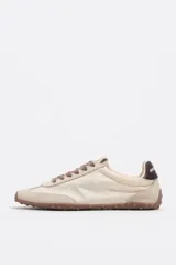 Zapatillas deportivas de color beige y crudo, confeccionadas en serraje vacuno con inserciones de tela, cordones marrones y suela de caucho con tacos. Cuenta con detalle de piel marrón en la parte trasera con logo.