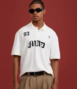 Remera de corte boxy confeccionada en algodón, con cuello polo y cierre frontal de botones. Presenta un diseño estampado en el frente con el número 93, una cruz y la palabra MAD en tipografía gótica.