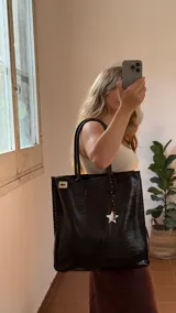 Tote bag de ecocuero color marrón oscuro con textura de cocodrilo, detalles de tachas en los laterales y asas de cuero marrón liso. Incluye un dije de guampa blanca colgando.