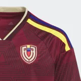 Camiseta de fútbol Adidas, modelo Local 2026 para fanáticos, color granate con patrón geométrico de triángulos en tono más oscuro. Presenta cuello en V con ribete beige y detalles en amarillo y azul en los hombros. Lleva el escudo de la selección venezolana y el logo de Adidas bordados.
