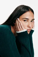 Saco corto de punto trenzado fino en color verde oscuro, con cuello redondo, cierre frontal de botones y bolsillos delanteros. Presenta detalles de ribetes festoneados en color crema en los bolsillos y en los puños.