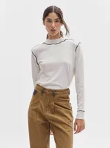 Polera de cuello alto confeccionada en tela rib color blanco, con mangas largas y hombreras. Presenta pespuntes en contraste en las terminaciones del cuello, hombros y puños.