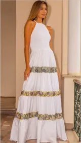 Vestido largo blanco con escote halter y cuerpo superior fruncido. La falda presenta un diseño escalonado con detalles de bordados florales en tonos azules y verdes en cada nivel, finalizando con un borde inferior decorado con pequeños pompones.
