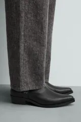 Bota de media caña en color negro, con diseño de arnés que incluye tiras, argollas metálicas y tachas decorativas alrededor del tobillo. Presenta punta fina y tacón cuadrado.