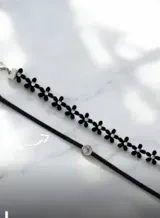 Gargantilla doble estilo choker, compuesta por una tira de flores negras con centro de cristal y una tira de gamuza negra con un dije circular de cristal central.
