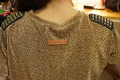 Vestido corto tipo remerón de lycra con acabado brillante color dorado. Presenta cuello redondo, mangas cortas y detalle de hombreras decoradas con tachas y cadenas metálicas.