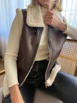 Chaleco de gamulán color marrón con interior y bordes de corderito sintético color crema. Presenta cierre frontal metálico y diseño sin mangas, ideal para superponer sobre otras prendas.