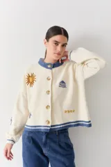 Cardigan de lana color crema con cuello redondo, cierre frontal de botones y mangas largas con puños ajustados. Presenta bordados decorativos en el frente que incluyen un sol, olas y estrellas, con detalles en azul en el cuello y el ruedo.
