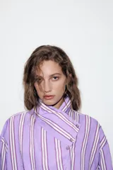 Camisa de corte oversize con diseño de rayas verticales en tonos violeta, blanco y bordó. Presenta cuello subido cruzado con cierre de botones frontal, mangas largas con puños y bajo asimétrico.
